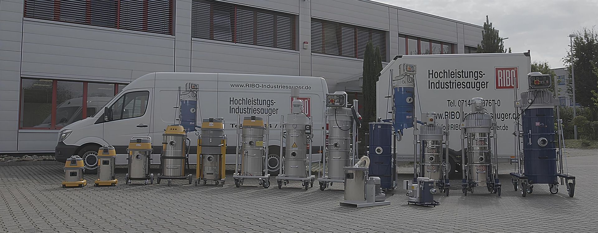 Mehrere industrielle Hochleistungsstaubsauger verschiedener Bauarten stehen vor einem Gebäude mit zwei Transportern im Hintergrund